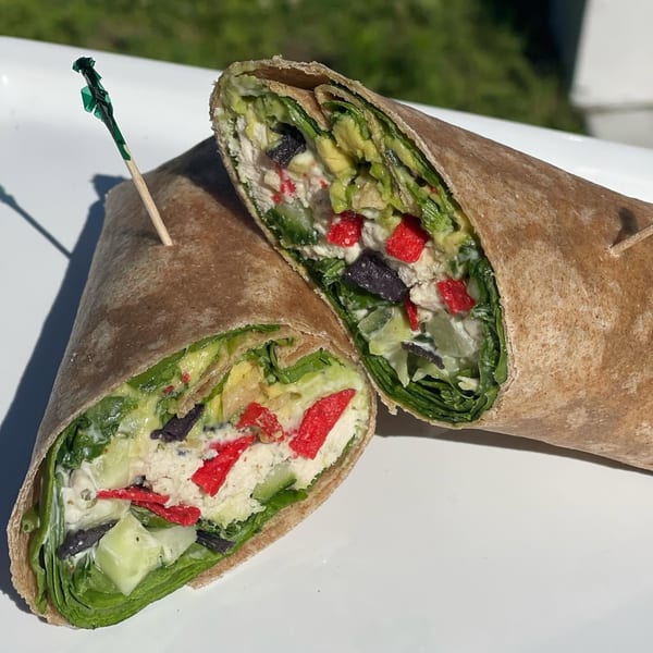 Two cut whole-wheat wraps filled with leafy greens, avocado, creamy spread, cucumber, red peppers, and dark beans on a white table.