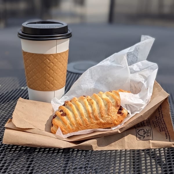 Takeaway coffee cup and chocolate croissant on a metal outdoor table in sunlight.