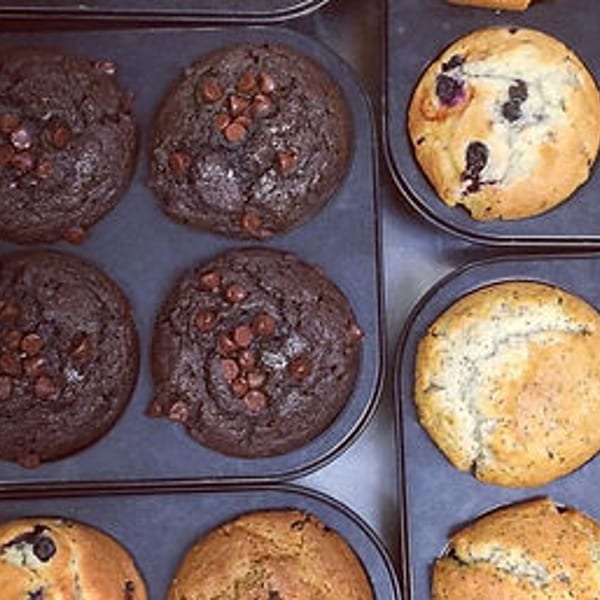 Trays of freshly baked muffins, including chocolate chip, blueberry, and plain varieties.