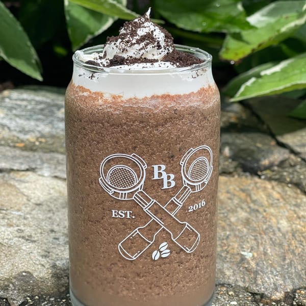 Chocolate blended drink topped with whipped cream and cookie crumbs in a branded glass, set outdoors among green leaves.