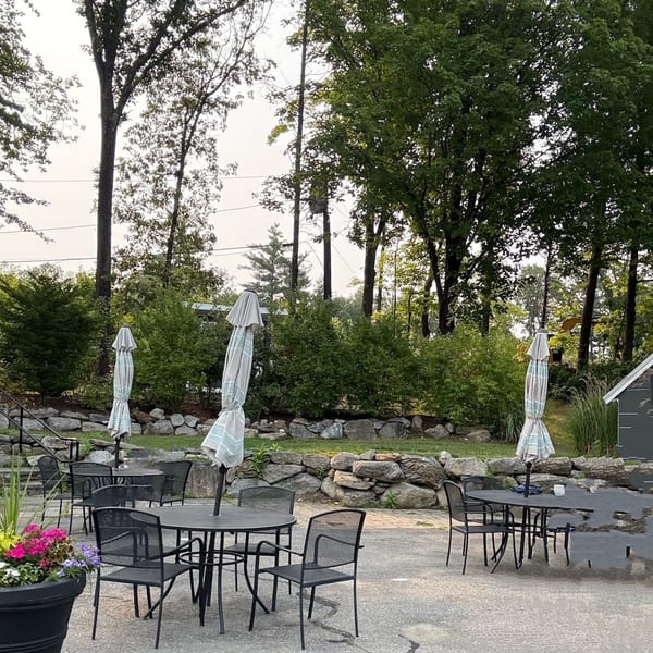 Outdoor patio with black tables and chairs, striped umbrellas, and lush green trees and bushes.