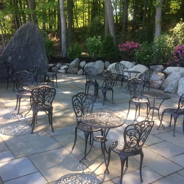 An outdoor patio with ornate black metal tables and chairs, surrounded by rocks, trees, and pink flowers.