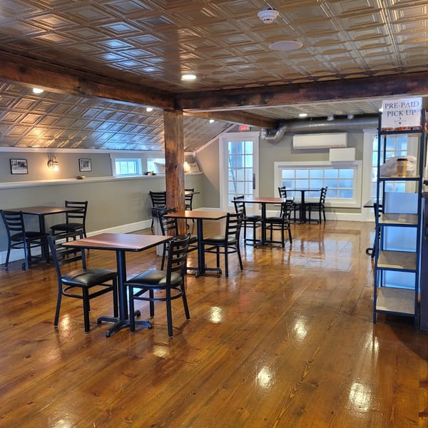 Empty restaurant dining room with wooden floors, black tables and chairs, and a patterned ceiling.